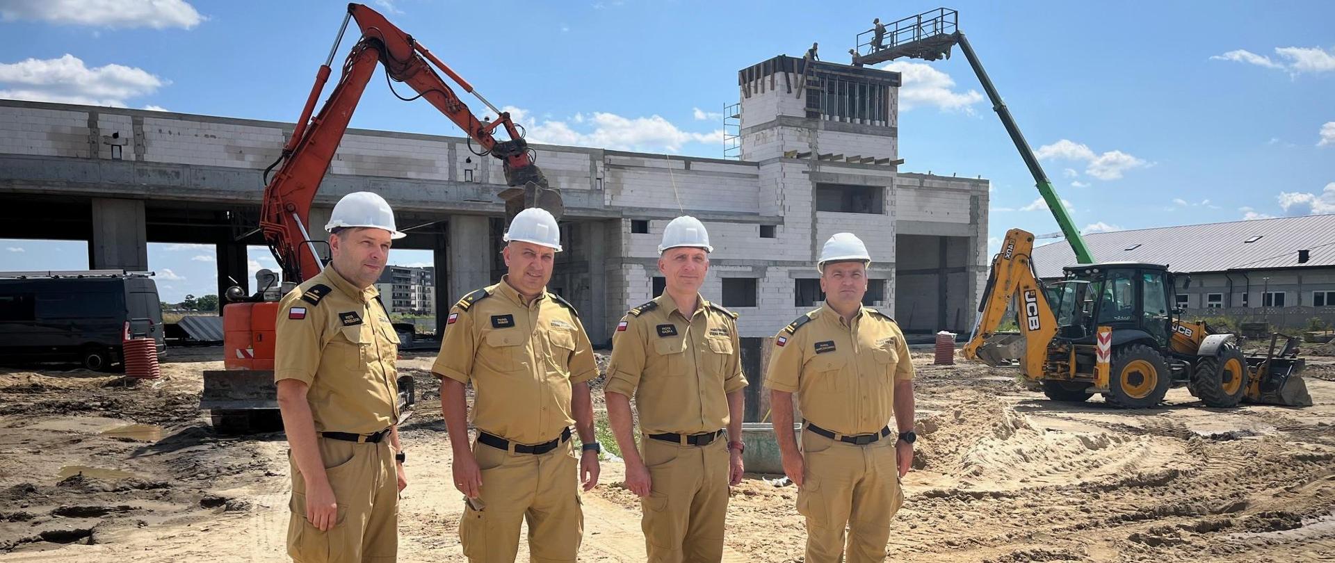 Czterej strażacy w piaskowym umundurowaniu, noszący na głowach białe kaski. W tle budowa obiektu