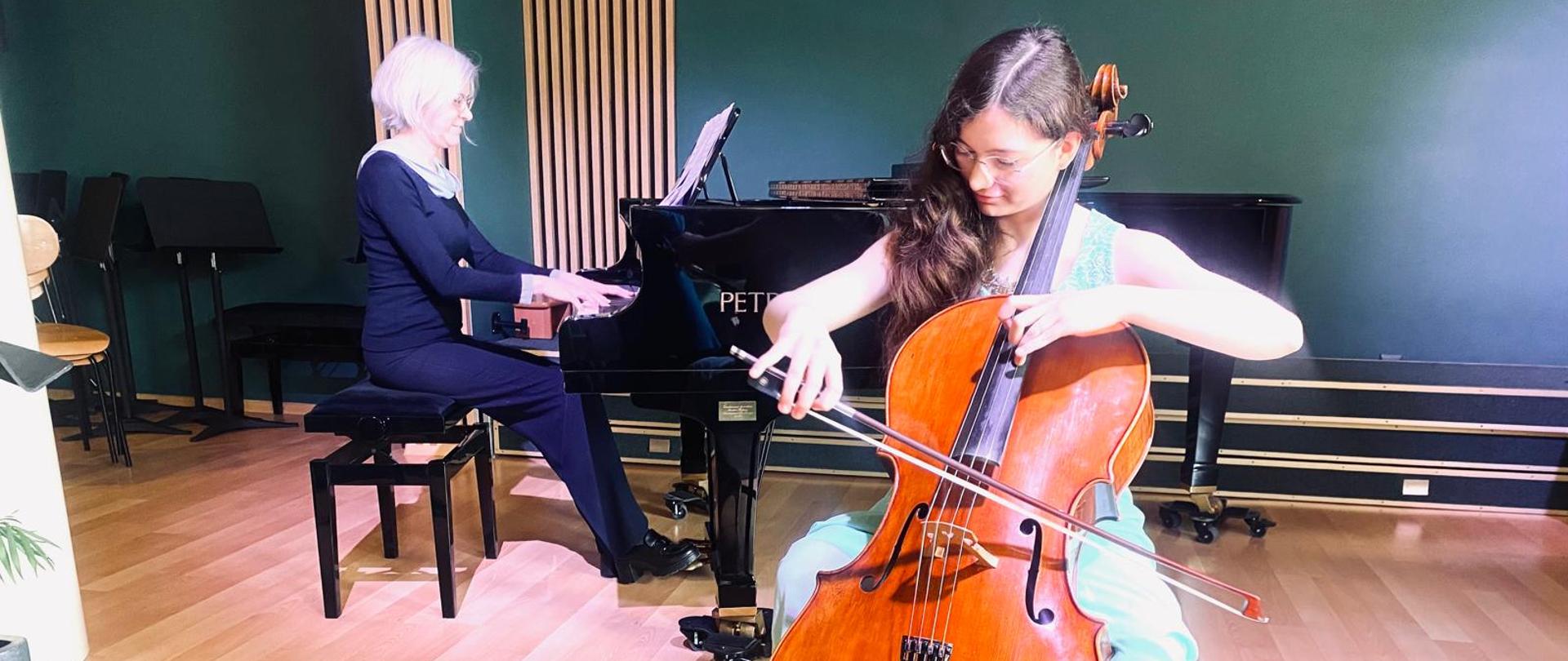 Na zdjęciu widać dwie kobiety grające razem muzykę kameralną. Na pierwszym planie, z prawej strony, młoda kobieta w jasnoniebieskiej sukni gra na wiolonczeli, skupiona na instrumencie. W tle, po lewej stronie, starsza kobieta o siwych włosach, ubrana na ciemno, gra na fortepianie koncertowym marki Petrof. Obie artystki znajdują się w eleganckim, nowocześnie urządzonym pomieszczeniu z drewnianą podłogą i akustycznymi panelami na ścianie. Scena oświetlona jest ciepłym światłem, które podkreśla atmosferę skupienia i harmonii.