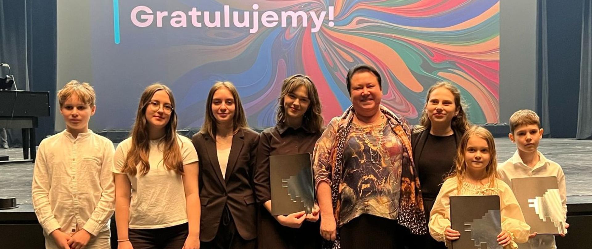 Widzimy grupę złożoną z młodzieży szkolnej. Wszyscy uśmiechnięci, niektórzy z nich trzymają przyznane im dyplomy z przyznaniem nagrody. W tle ogromna scena białostockiej opery i duży napis "Gratulujemy!" na kolorowym tle.