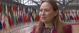 Minister funduszy i polityki regionalnej Katarzyna Pełczyńska-Nałęcz stoi w korytarzu i mówi do mikrofonu dziennikarskiego, za jej plecami flagi państw członkowskich UE