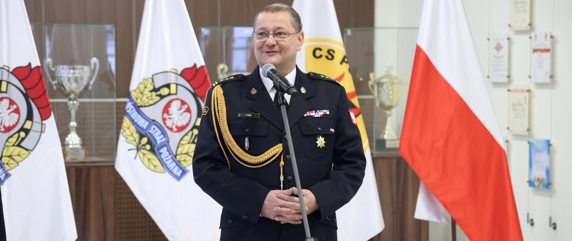 Odchodzący na emeryturę Zastępca Komendanta Centralnej Szkoły PSP przemawiający do mikrofonu, w tle flagi stojące na stojakach