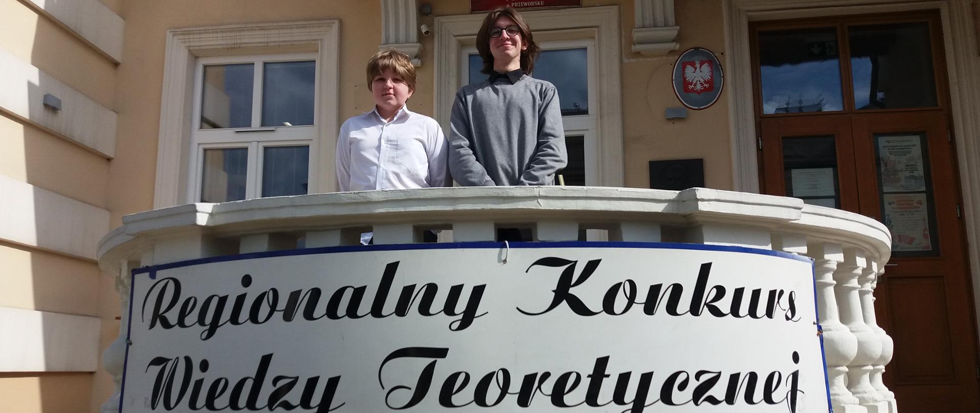 Na zdjęciu widnieją uczniowie PSM I stopnia w Jaśle, Hubert Michałowski i Jakub Michałowski,
którzy stoją na tarasie Państwowej Szkoły Muzycznej I stopnia w Przeworsku. Na balustradzie
widnieje napis: Regionalny Konkurs Wiedzy Teoretycznej. Kolorystyka zdjęcia jest beżowo-biało -
czarno-niebieska.