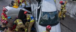 Strażacy PSP prowadzą działania ratownictwa technicznego przy dwóch uszkodzonych samochodach podczas zawodów, a przebieg akcji z góry obserwują sędziowie oraz zgromadzeni widzowie.

