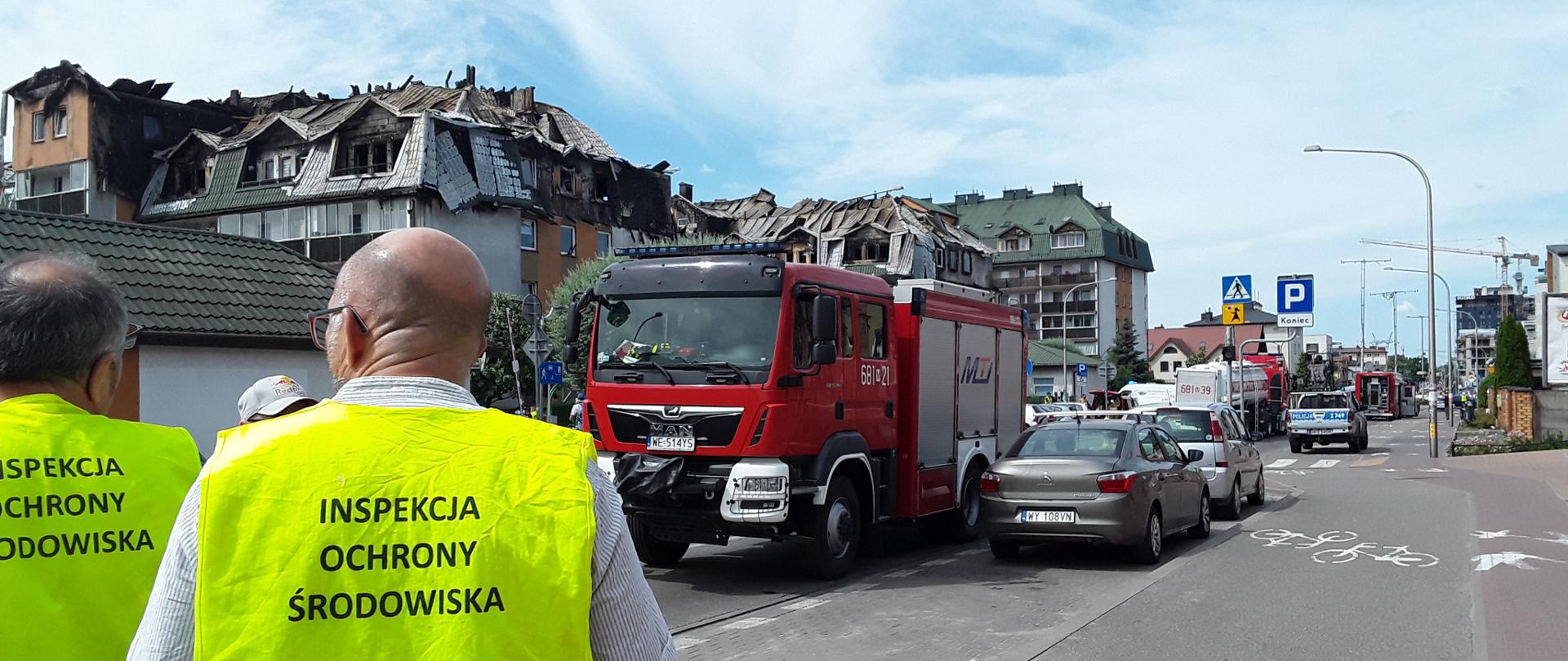 Dwóch inspektorów Mazowieckiego Wojewódzkiego Inspektoratu Ochrony Środowiska prowadzi działania w terenie zabudowanym. W tle widać wóz bojowy straży pożarnej stojący na asfaltowej drodze oraz kilka samochodów osobowych. Za nim stoi budynek z uszkodzonym przez pożar dachem.