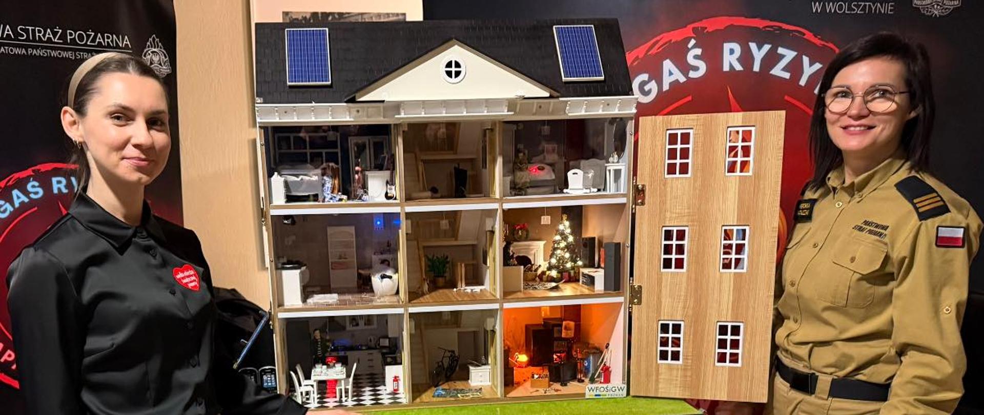 Na zdjęciu widoczny jest strażak Państwowej Straży Pożarnej stojący w pomieszczeniu wystawowym. Po lewej stronie znajduje się duży, otwarty model domu pokazujący wnętrza pomieszczeń mieszkalnych – kuchnię, salon i pokoje, z elementami instalacji oraz wyposażenia. Za strażakiem widoczna jest czarna ścianka z napisem „Państwowa Straż Pożarna – Komenda Powiatowa PSP w Wolsztynie” oraz hasłem „Zgaś ryzyko”. Po prawej stronie stoją stoliki z rozłożonym sprzętem edukacyjnym i ratowniczym. Całość ma charakter stanowiska informacyjno-edukacyjnego.