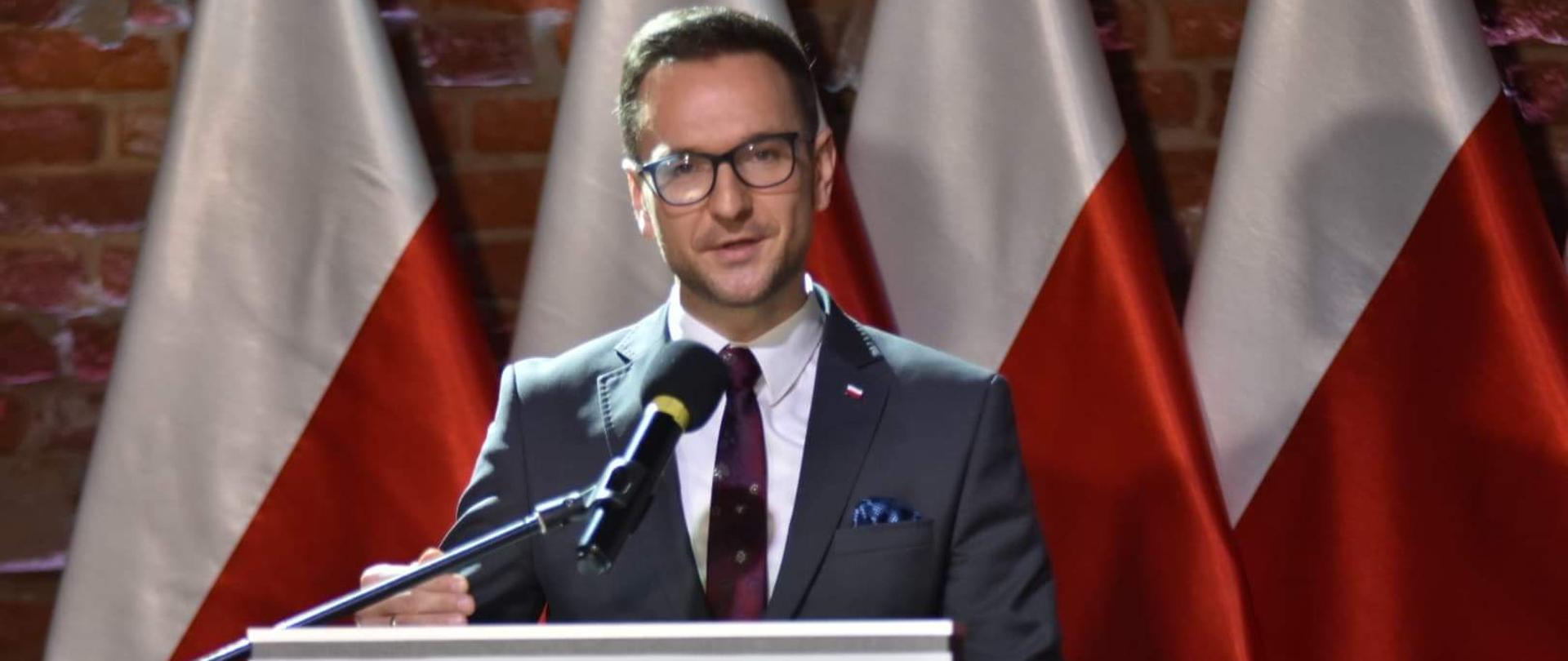 Na zdjęciu wiceminister Waldemar Buda stoi przy mównicy, w tle cztery biało-czerwone flagi.
