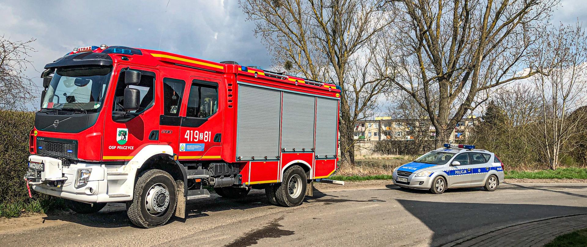 Na drodze znajduje się wóz strażacki OSP Pełczyce, a za nim stoi radiowóz. W oddali znajdują się drzewa oraz blok mieszkalny. Zdjęcie wykonano w porze dziennej.