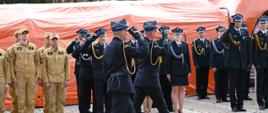 Zdjęcie przedstawiające grupę strażaków na wybrukowanym placu przed dużym, pomarańczowym namiotem. Na pierwszym planie widać dwóch oficerów w czarnych mundurach galowych, którzy maszerują i oddają honor, salutując. W tle, po lewej stronie, stoi grupa strażaków w piaskowych mundurach służbowych, natomiast po prawej i w głębi ustawiony jest szereg funkcjonariuszy w galowych mundurach z czapkami rogatywkami. Atmosfera jest podniosła i oficjalna.