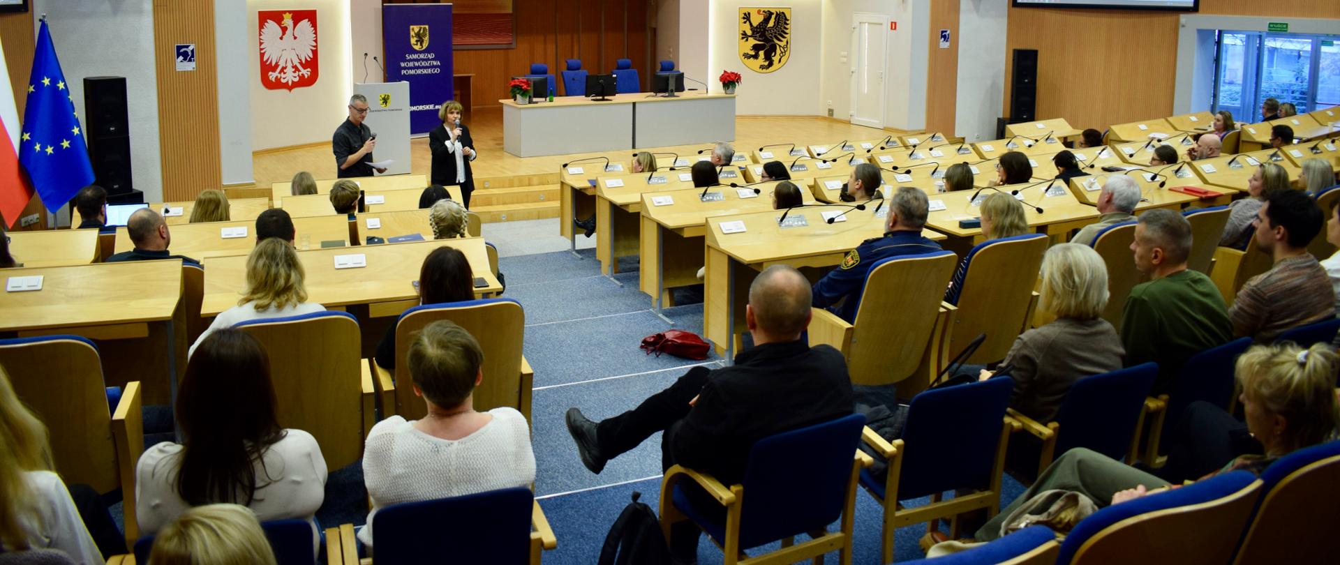 kilkanaście osób siedzi w dużej sali i słucha prelegenta