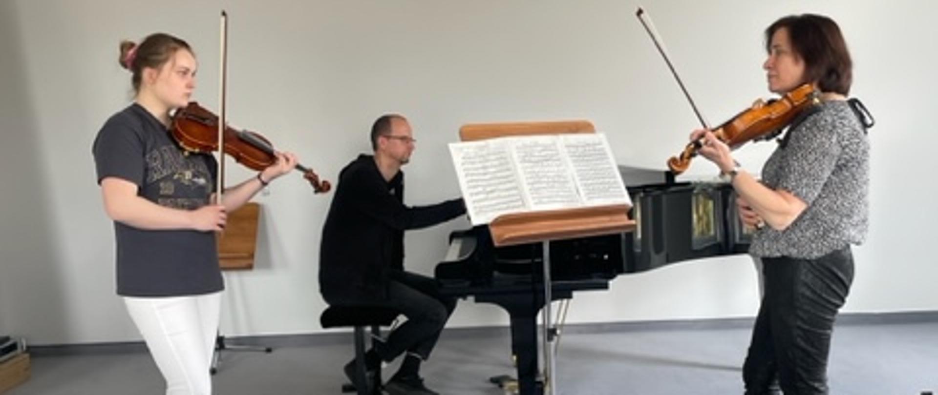 Na sali kameralnej uczennica gra na skrzypcach na przeciw prowadzącej warsztaty. Pośrodku nich pulpit a na nim nuty. Na pierwszym planie cztery puste krzesła. Z tyłu fortepian, przy którym siedzi akompaniator