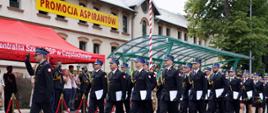 Zdjęcie przedstawia uroczysty przemarsz grupy strażaków w czarnych mundurach wyjściowych. Defilada odbywa się przed budynkiem Centralnej Szkoły Państwowej Straży Pożarnej w Częstochowie, co potwierdza napis na czerwonym namiocie z lewej strony zdjęcia. Nad wejściem do budynku widnieje duży, żółty transparent z czerwonym napisem: „PROMOCJA ASPIRANTÓW”. Strażacy maszerują w szyku, ubrani są w czarne mundury z czapkami rogatywkami z niebieskim otokiem, niektórzy z nich noszą również białe rękawiczki i złote sznury na ramieniu. W rękach trzymają dokumenty awansu. Na chodniku po lewej stronie stoją osoby obserwujące i fotografujące przemarsz — część z nich to również strażacy w galowych mundurach. W tle widać budynek szkoły oraz zieloną metalową konstrukcję z daszkiem.