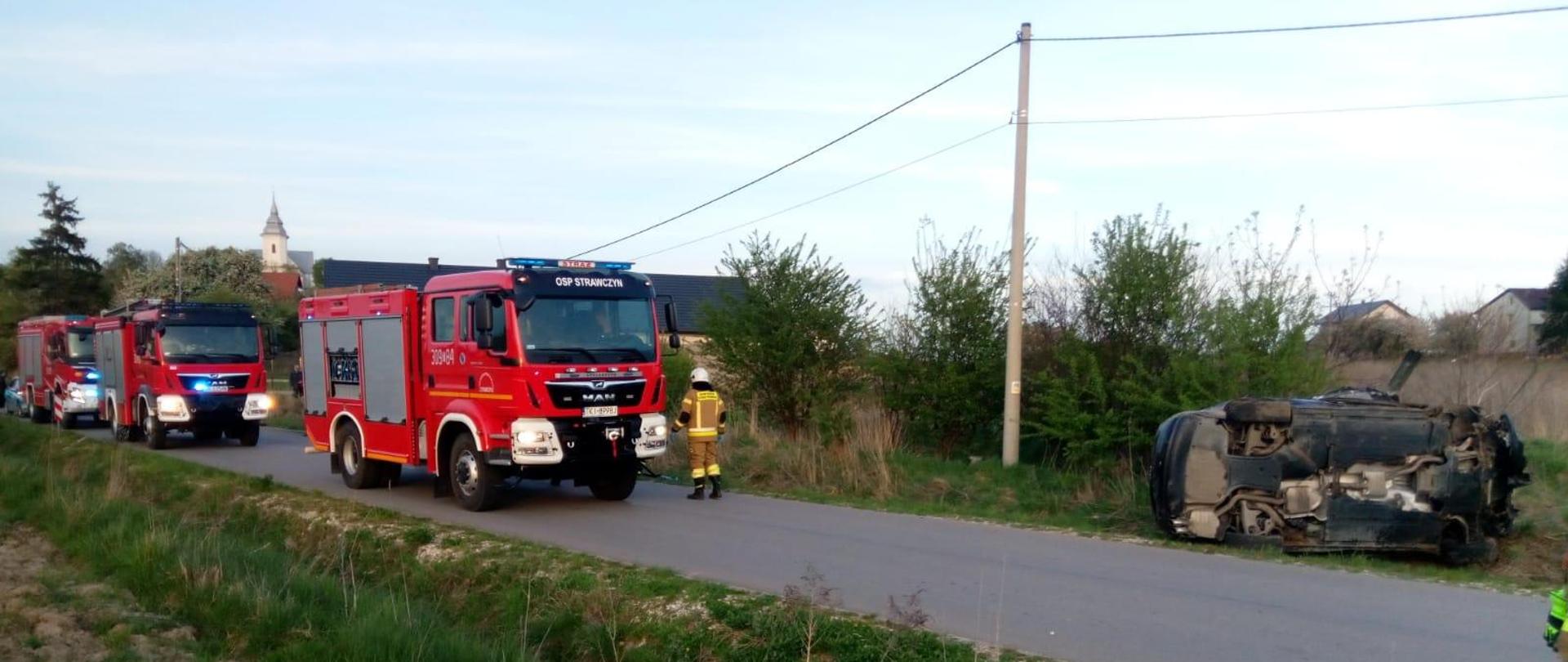 Droga powiatowa: po prawej stronie leżący na poboczu w pozycji na boku samochód osobowy. Po lewej stronie sznur samochodów służb ratowniczych. 