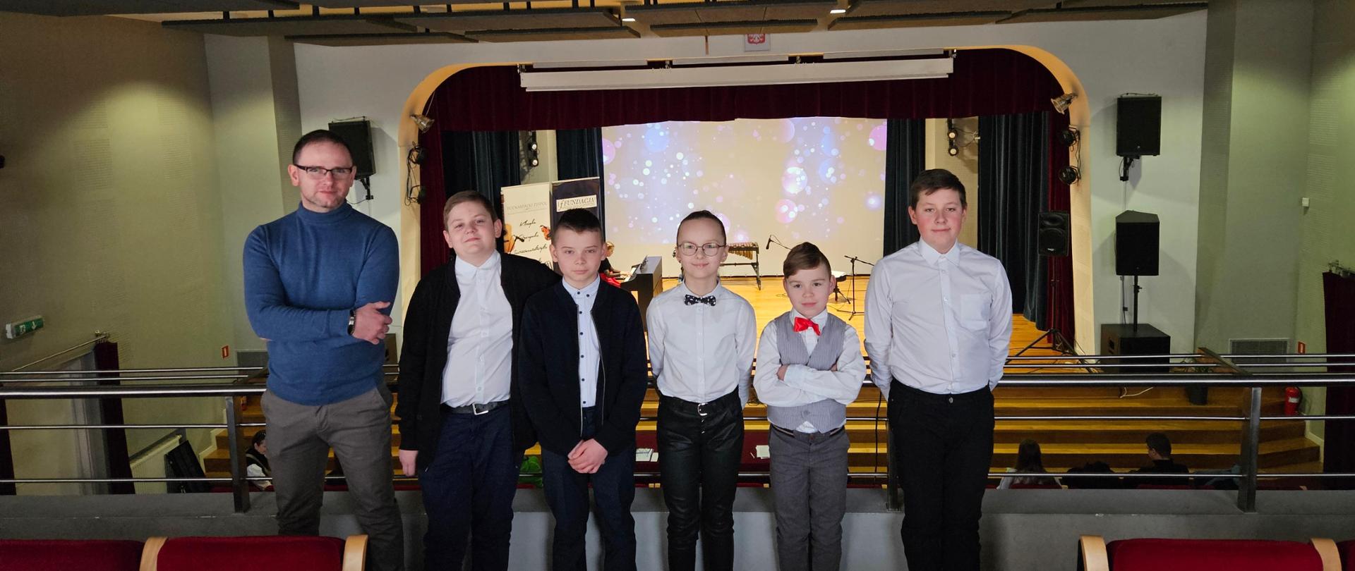 Zdjęcie uczniów oraz ich nauczyciela - zdj. wykonane na balkonie sali koncertowej, w tle widoczna scena konkursowa