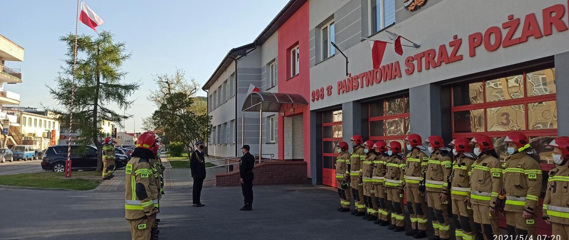 Na placu przed budynkiem w dwóch szeregach stoją strażacy w umundurowaniu specjalnym . Przed szeregiem widoczni dwaj strażacy zwróceni do siebie przodem, stoją w niewielkiej odległości od siebie. Jeden ze strażaków występuje w umundurowaniu wyjściowym z czapką rogatywką i sznurem galowym, natomiast drugi w umundurowaniu dowódczym. Przed budynkiem, w oddali na maszcie powiewa zawieszona flaga Rzeczypospolitej Polskiej. Przed masztem stoi trzech strażaków. 