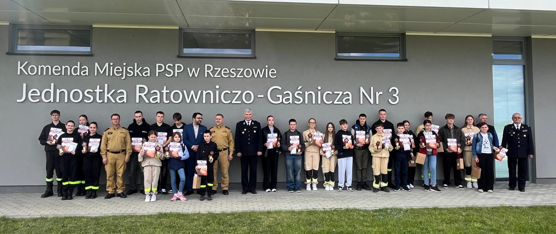 Duża grupa uczestników konkursu, opiekunów oraz strażaków pozuje do wspólnego zdjęcia przed budynkiem Komendy Miejskiej PSP w Rzeszowie – Jednostki Ratowniczo-Gaśniczej nr 3. Uczestnicy trzymają dyplomy i nagrody. Na elewacji budynku widoczny jest napis z nazwą jednostki. Zdjęcie wykonano na zewnątrz, na tle nowoczesnego budynku straży pożarnej.