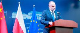Ambasador Jakub Kumoch wygaszający przemówienie na tle trzech flag