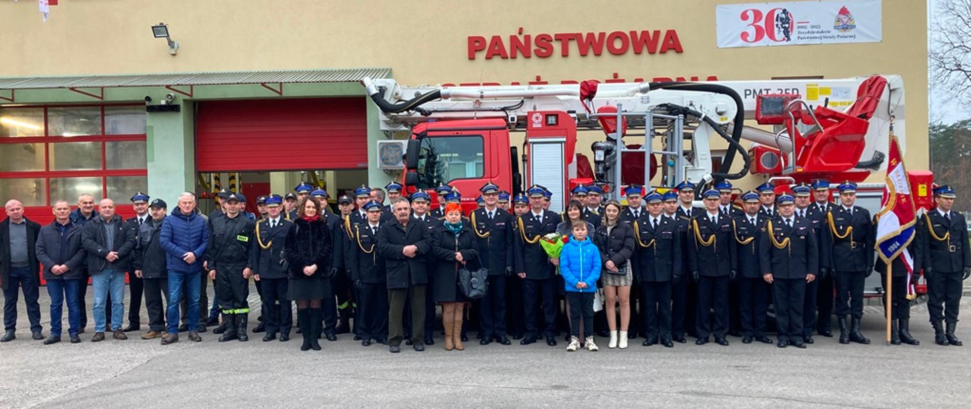 na zewnątrz, przed budynkiem remizy strażacy w luźnym szyku pozują do fotografii. 