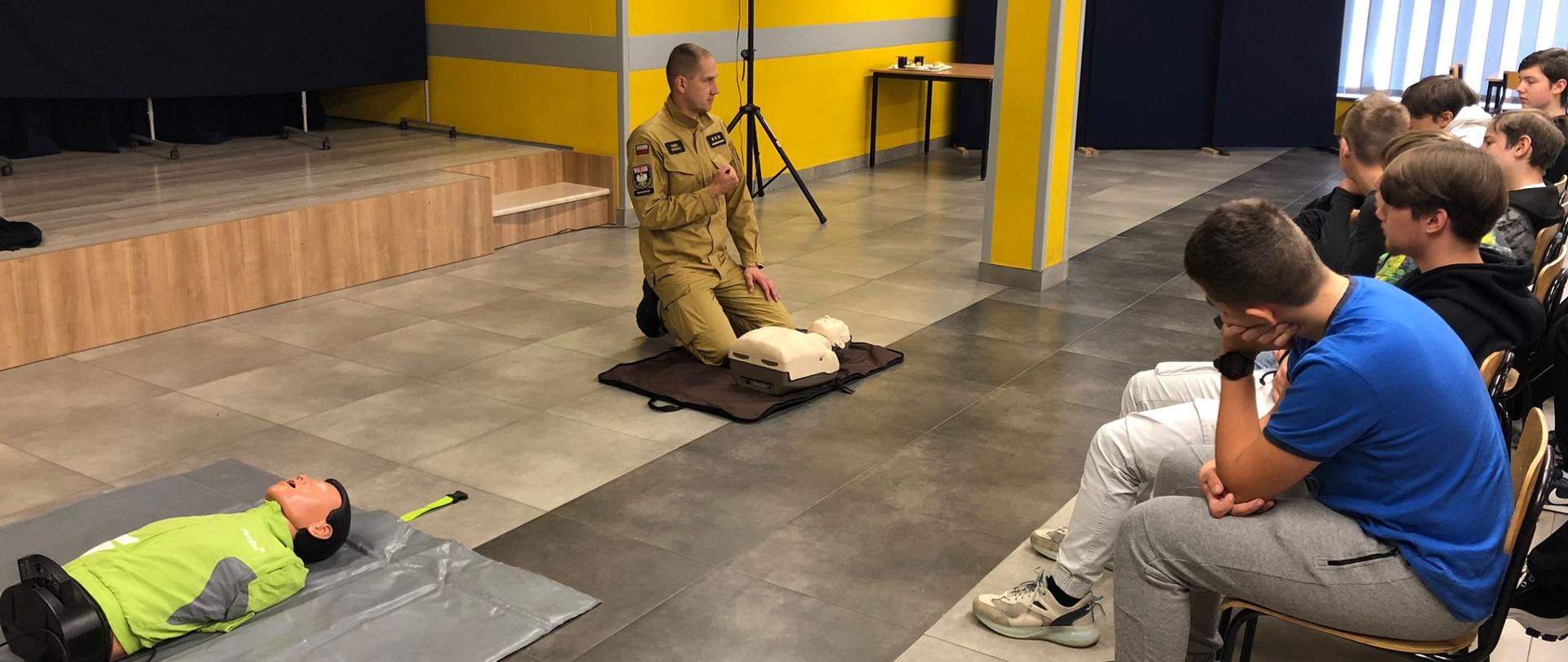 Widok z boku. Funkcjonariusz PSP klęczy na posadzce przy fantomie. Przed nim na stołkach siedzą uczniowie. Obok leży drugi, bardziej zaawansowany fantom. W tle żółta ściana i wyjście na podest zasłonięty granatową kotarą. 