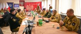 Strażacy ubrani w piaskowe mundury oraz białe kamizelki , część z nich siedzi przy stole reszta stoi obok. Na stołach postawione są laptopy oraz zielone butelki z wodą mineralną. W tle godło Polski