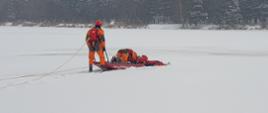 Na zdjęciu widzimy dwóch ratowników Państwowej Straży Pożarnej, którzy za pomocą specjalnych sań lodowych wciągnęli osobę, pod którą załamał się lód. Ratownicy, którzy pozostali na brzegu ściągają ich ze zbiornika wodnego za pomocą linek. Na szczęście był to tylko pokaz działań ratowniczych. Osobą poszkodowaną był bardzo dobrze wyszkolony ratownik, zabezpieczony w specjalne ubranie wypornościowe, które nie pozwoliłoby mu utonąć. Był również zabezpieczony linką ratowniczą, która była przywiązana do strażackiego pasa. Cała symulacja zdarzenia odbyła się na zamarzniętym zbiorniku wodny, w taki sposób aby nikomu nic się nie stało