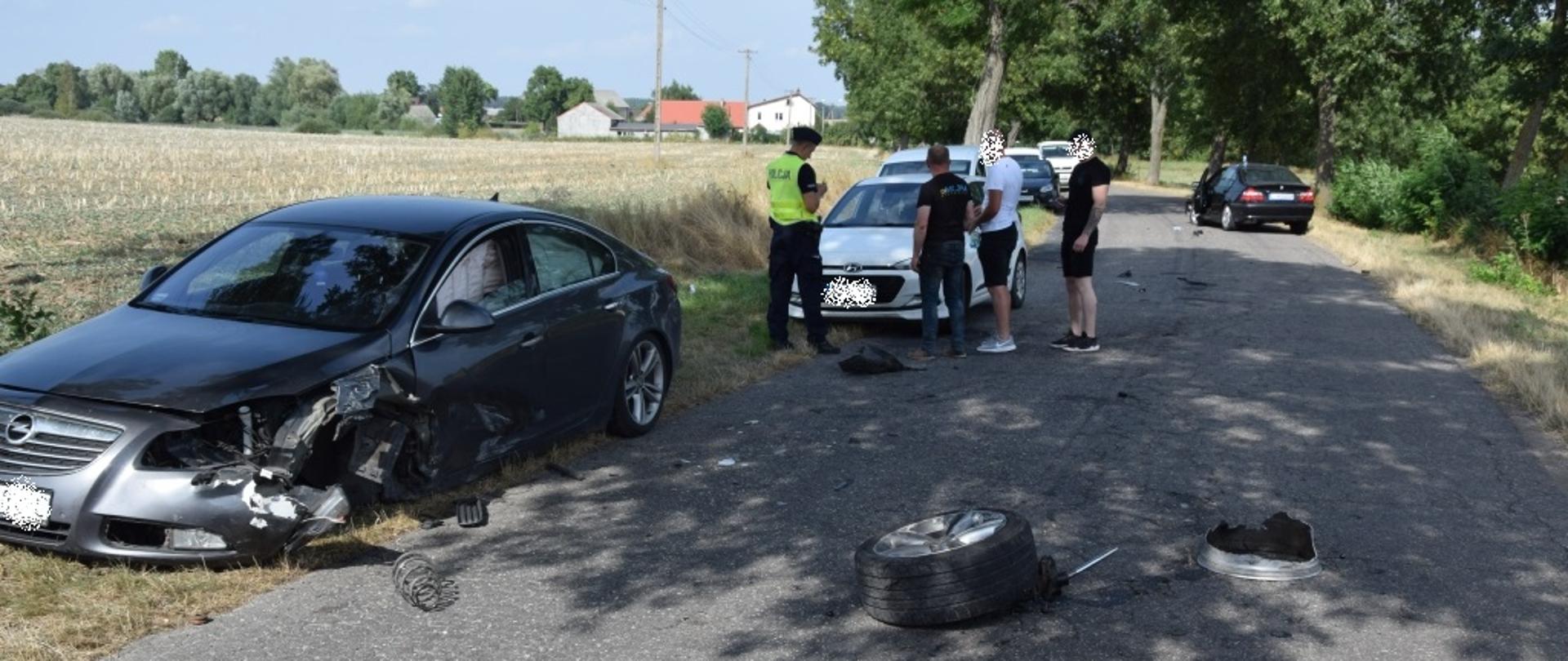Rozbite samochody osobowe. Na środku drogi leży oderwane koło. Po środku stoją 4 osoby, w tym policjanci. 