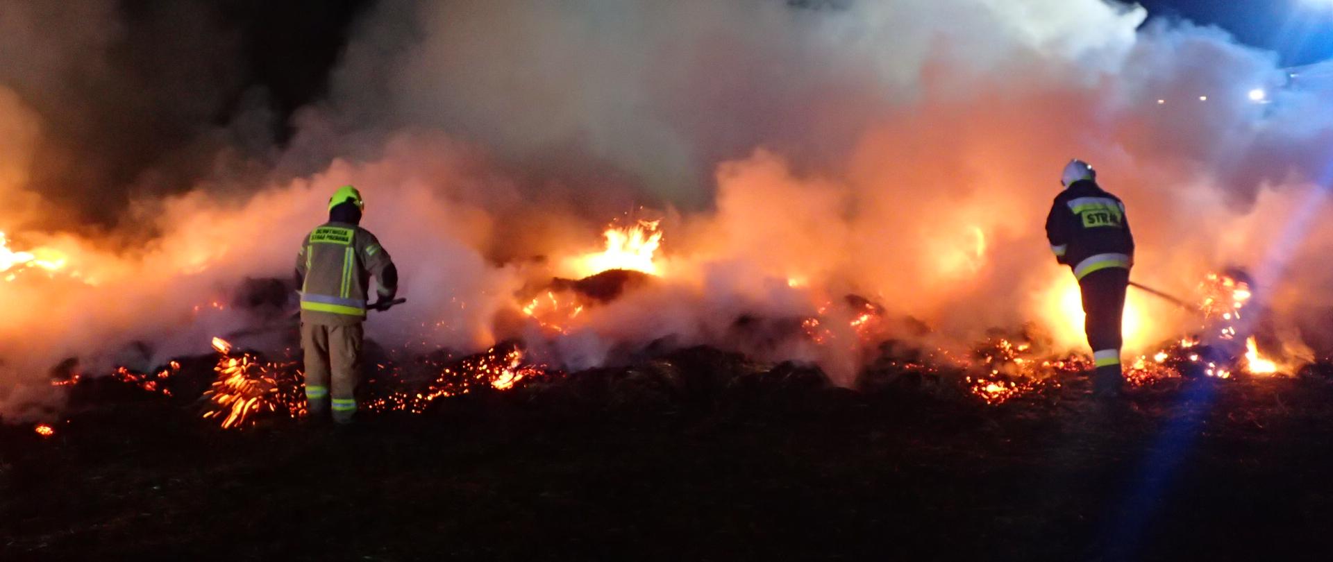 Strażacy OSP rozciągają płonące bele słomy. Dookoła dym i płomienie.