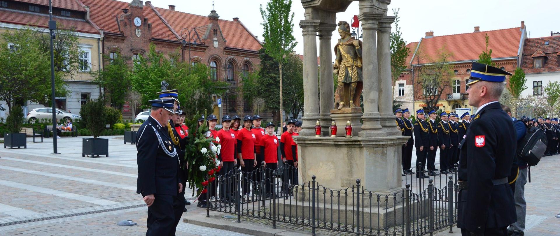 Zdjęcie przedstawia strażaków składających wiązankę pod pomnikiem św. Floriana w Myślenicach