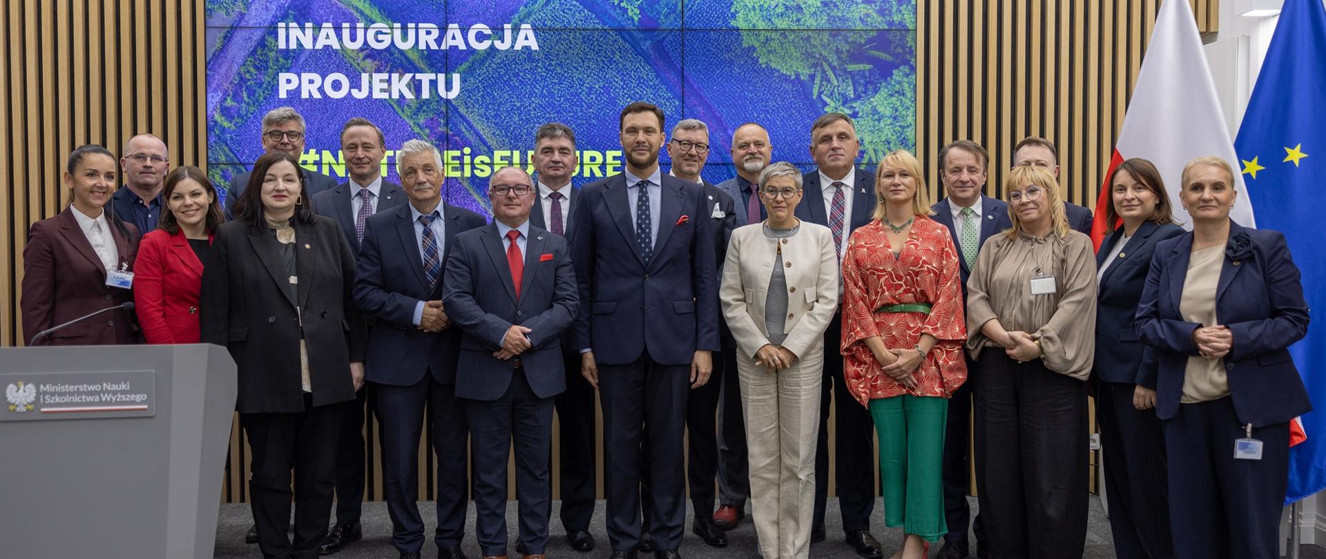 Zdjęcie zbiorowe - pod ścianą z cienkich pionowych listewek z wbudowanym wielkim ekranem z napisem Inauguracja projektu #NATUREisFUTURE stoi grupa dwudziestu osób, pośrodku wiceminister Mrówczyńska w białym garniturze.