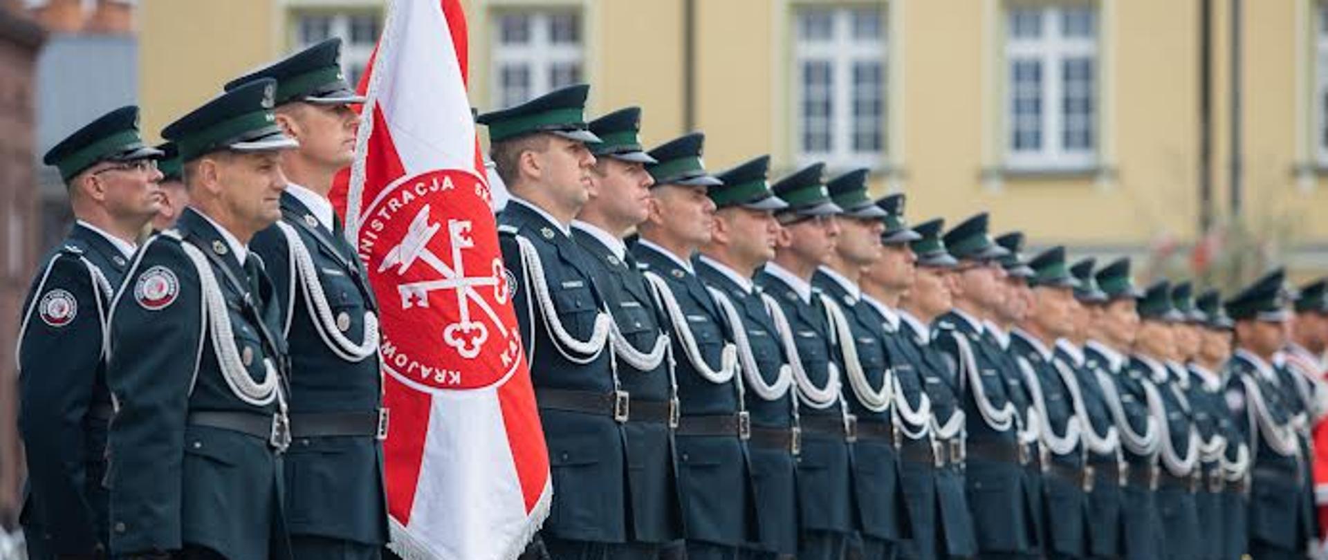 Kompania honorowa funkcjonariuszy KAS. Z przodu dowódca i poczet sztandarowy, dalej w szeregu pozostali członkowie kompanii (kilkadziesiąt funkcjonariuszy). Wszyscy w umundurowaniu galowym. W tle fragment kamienicy na Starym Rynku.