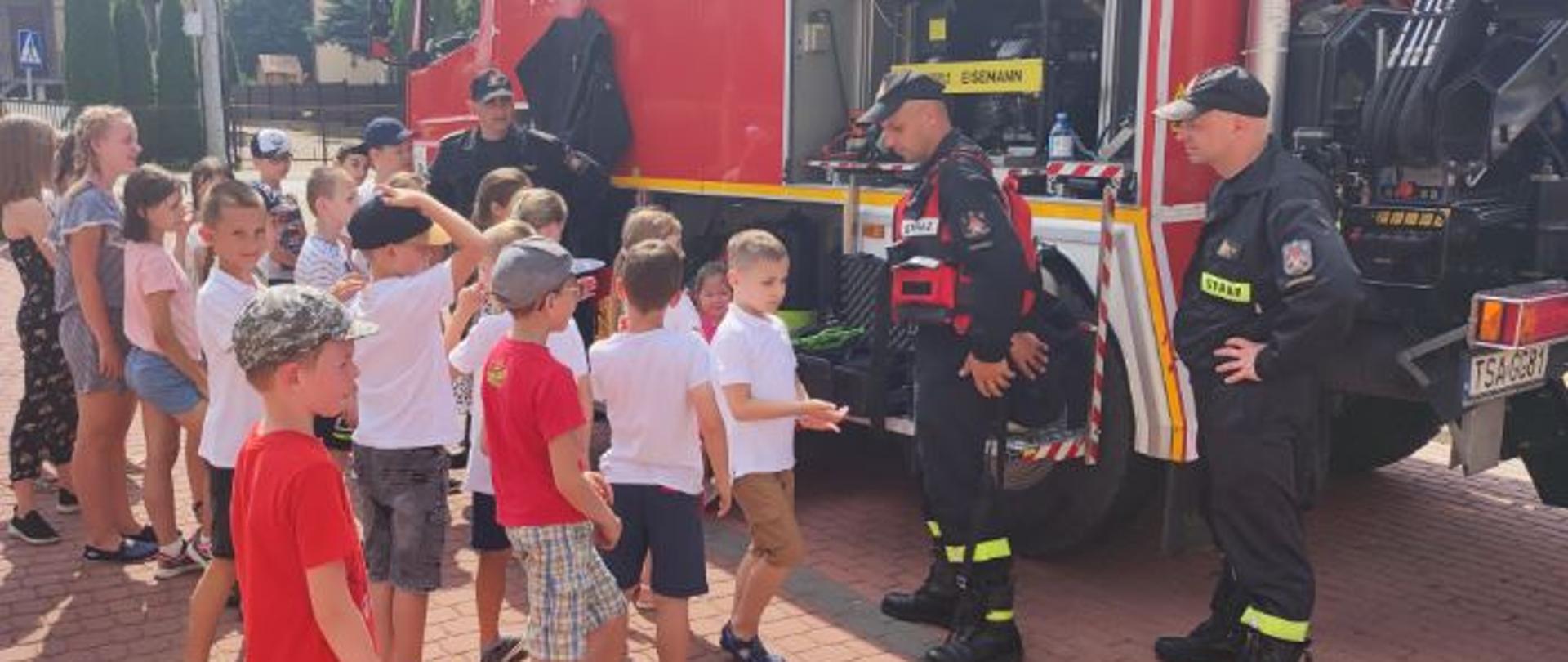 Zdjęcie zrobione w dzień. Na zdjęciu widać grupę młodzieży i trzech strażaków stojących na tle samochodu strażackiego przeznaczonego do ratownictwa wodnego. Jeden z strażaków ubrany jest w kamizelkę ratowniczą. 
