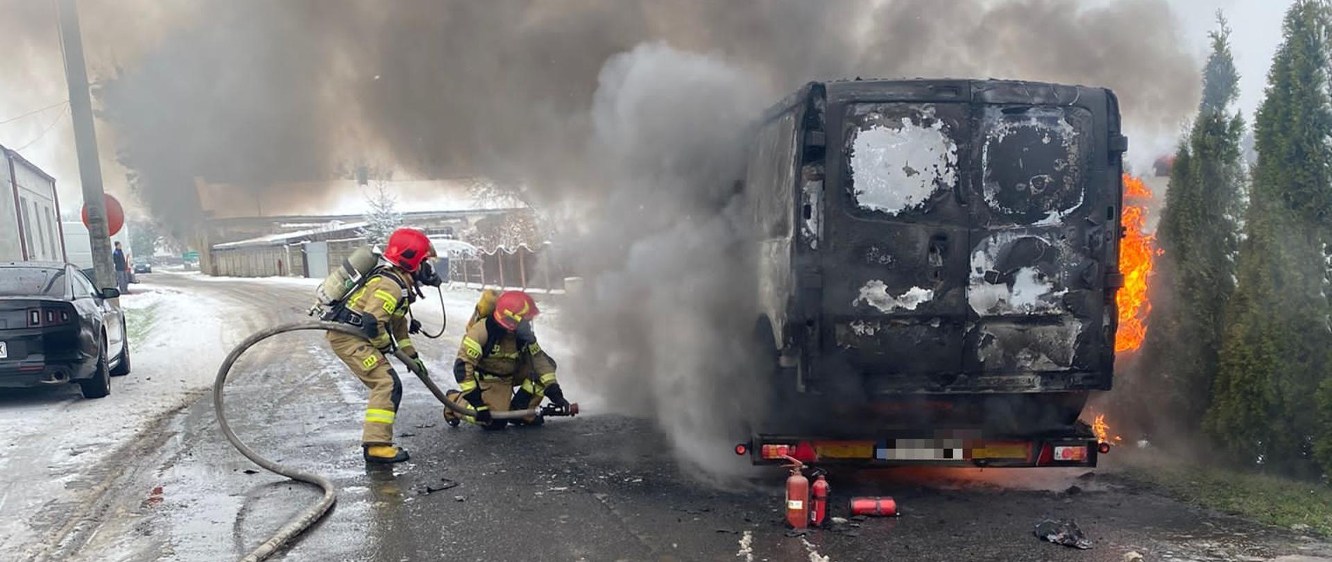 Na auto lawecie stoi płonący samochód typu bus przy nim dwóch strażaków gasi płonący samochód z boku widoczne płomienie oraz drzewka nad samochodem kłęby dymu