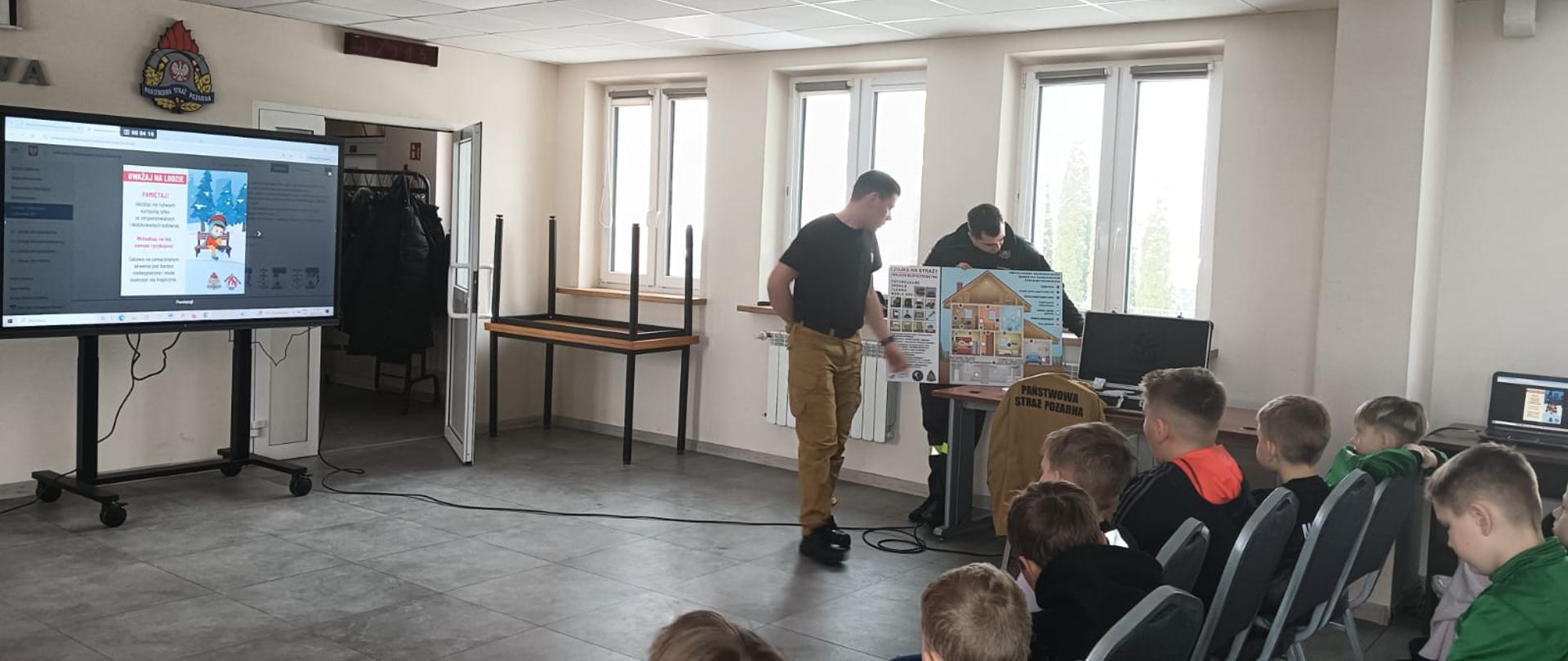 Dwóch funkcjonariuszy opowiada dzieciom o czujce dymu i czadu oraz ją prezentuje