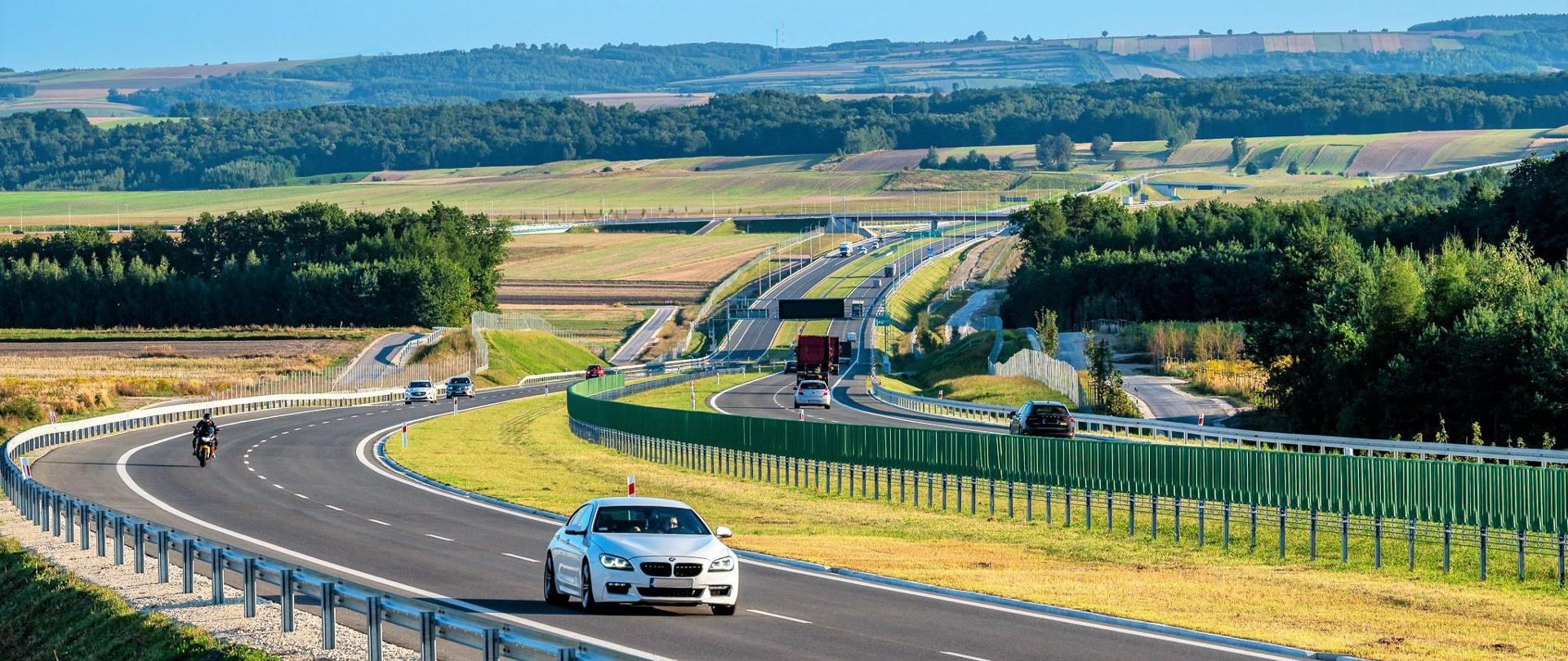 Droga ekspresowa S7 na odcinku Moczydło - Miechów. Dwujezdniowa droga ekspresowa wijąca się między polami i lasami po niewielkich pagórkach. Widoczne pojazdy poruszające się w obu kierunkach.