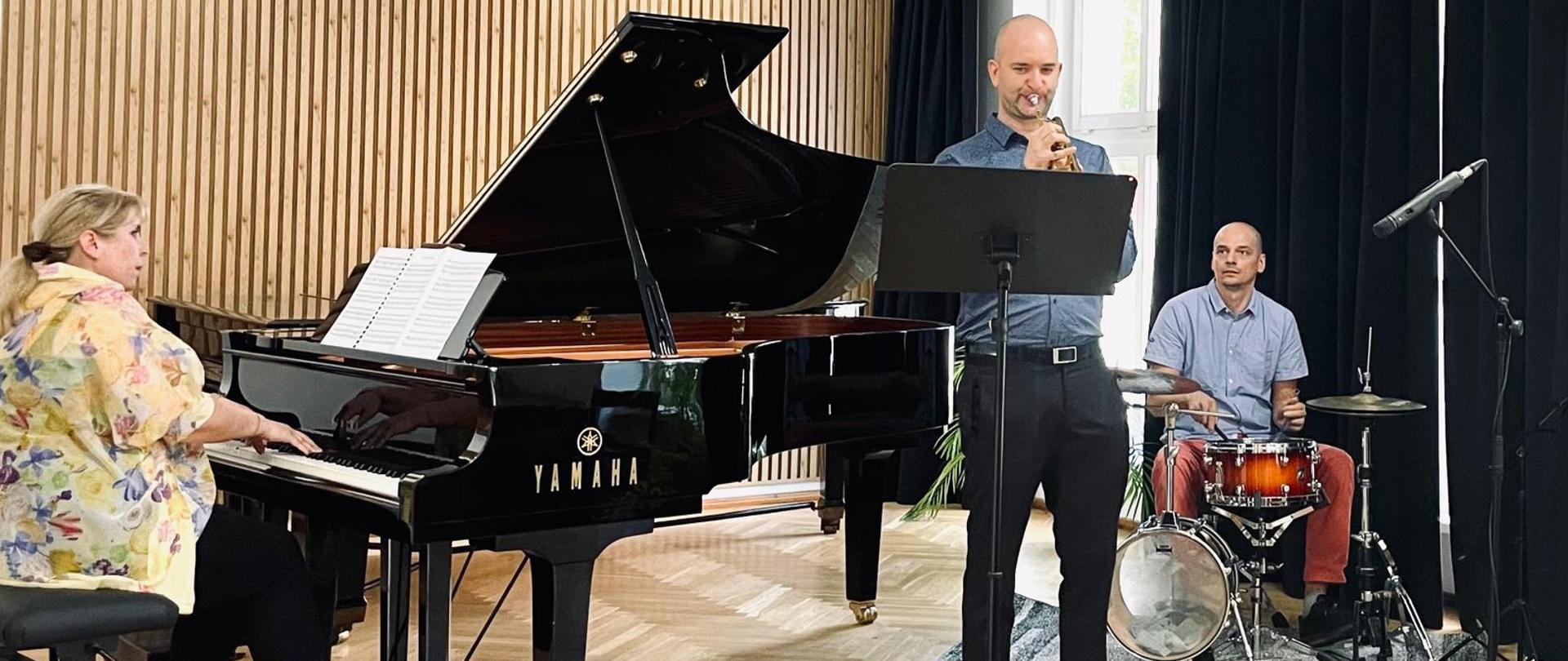 Nauczyciel klasy trąbki Wojciech Lizurej gra na trąbce, w tle po lewej stronie na fortepianie akompaniuje Iryna Yakymets, po prawej stronie na perkusji Andrzej Habdas, nauczyciele grają na instrumentach na scenie sali koncertowej PSM w Kłodzku z okazji rozpoczęcia roku szkolnego 2025-2026