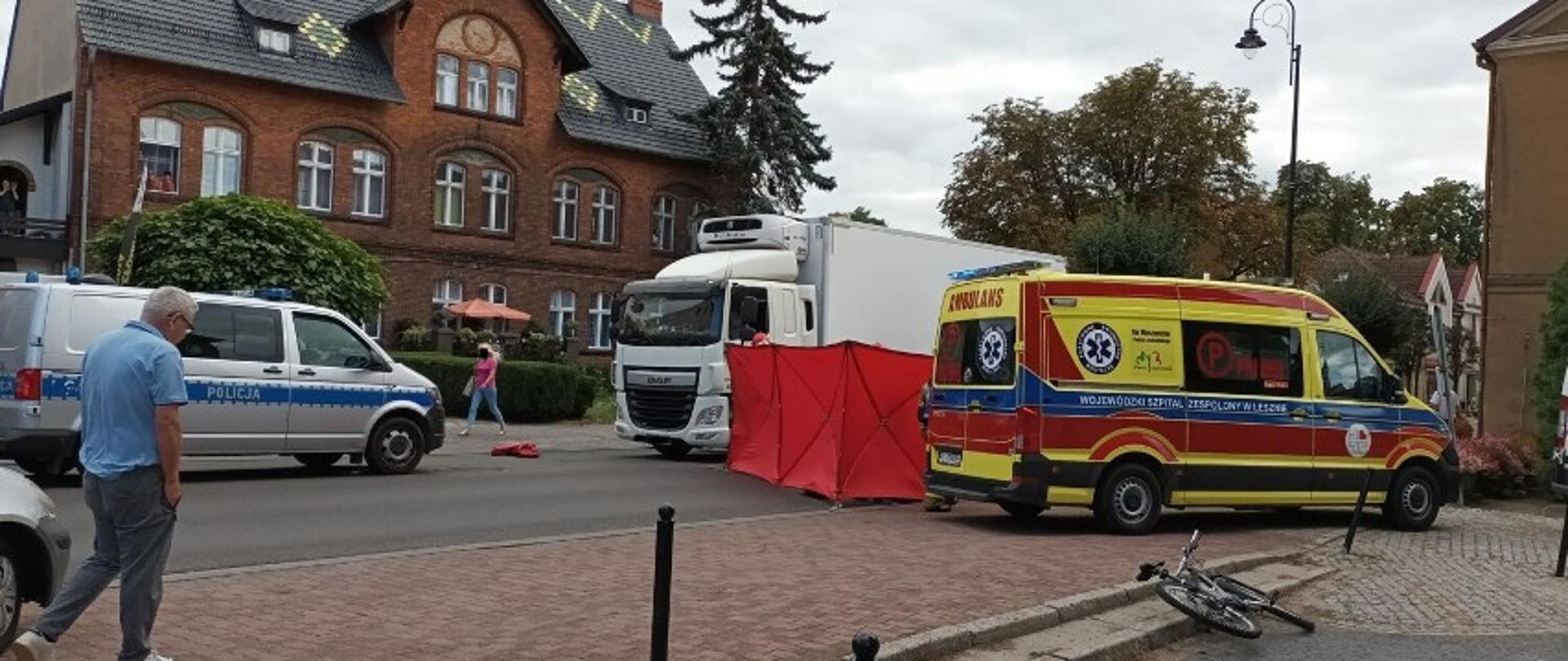 Zdjęcie wykonane na ulicy. Widoczny zespół karetki pogotowia, rozłożony parawan zasłaniający działania ratowników oraz samochód policji. Po ulicy chodzą przechodnie. W tle widać zabudowania.