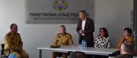 Na zdjęciu widoczna sala. Przy białych Stołach siedzą uczestnicy narady. Na ścianie w tle widoczne logo Państwowej Straży Pożarnej. Jedna z osób stoi i przemawia do reszty.