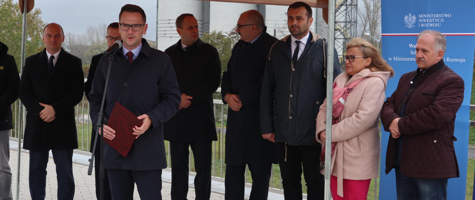 Pod wiatą stoi grupa ludzi. Obok baner ministerstwa. W pierwszym rzędzie do stojącego mikrofonu mówi wiceminister Waldemar Buda.
