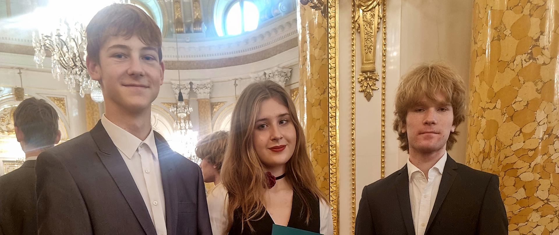 na zdjęciu sylwetki trzech osób dwóch męźczyzn w środku kobieta we wnętrzach pałacu królewskich białe ściany zdobione złotymi elementami