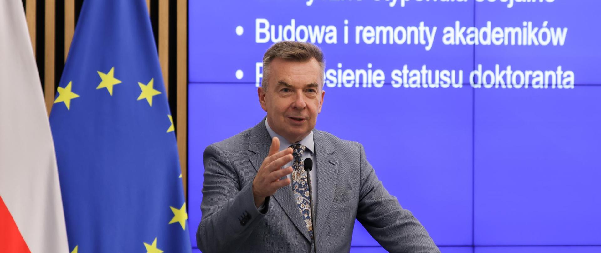 Minister Wieczorek stoi za mównicą i mówi do mikrofonu, za nim na wbudowanym w ścianę wielkim ekranie napis Elastyczne stypendia socjalne, budowa i remonty akademików, z boku flagi Polski i UE.