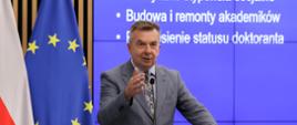 Minister Wieczorek stoi za mównicą i mówi do mikrofonu, za nim na wbudowanym w ścianę wielkim ekranie napis Elastyczne stypendia socjalne, budowa i remonty akademików, z boku flagi Polski i UE.