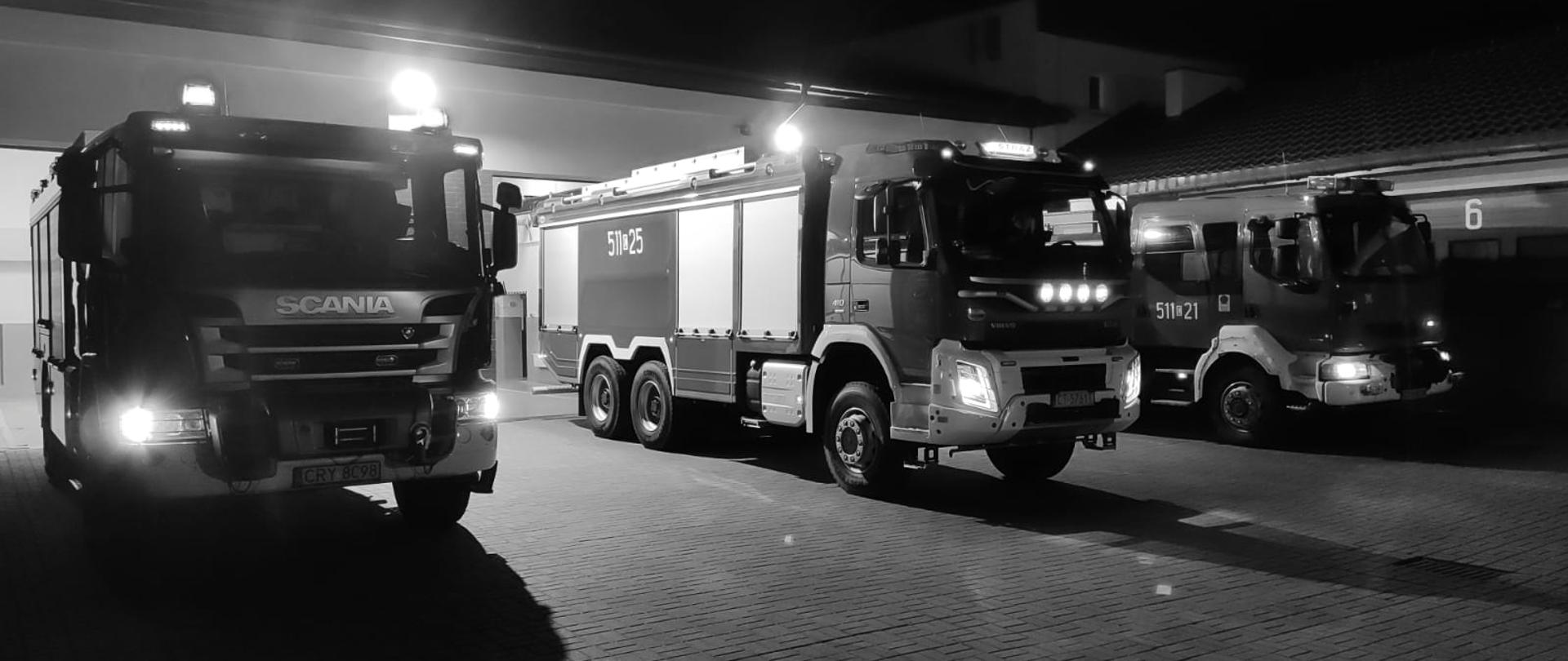 Zdjęcie przedstawia 3 samochody pożarnicze przed garażami. Samochody mają włączone światła alarmowe. Zdjęcie jest czaro białe.