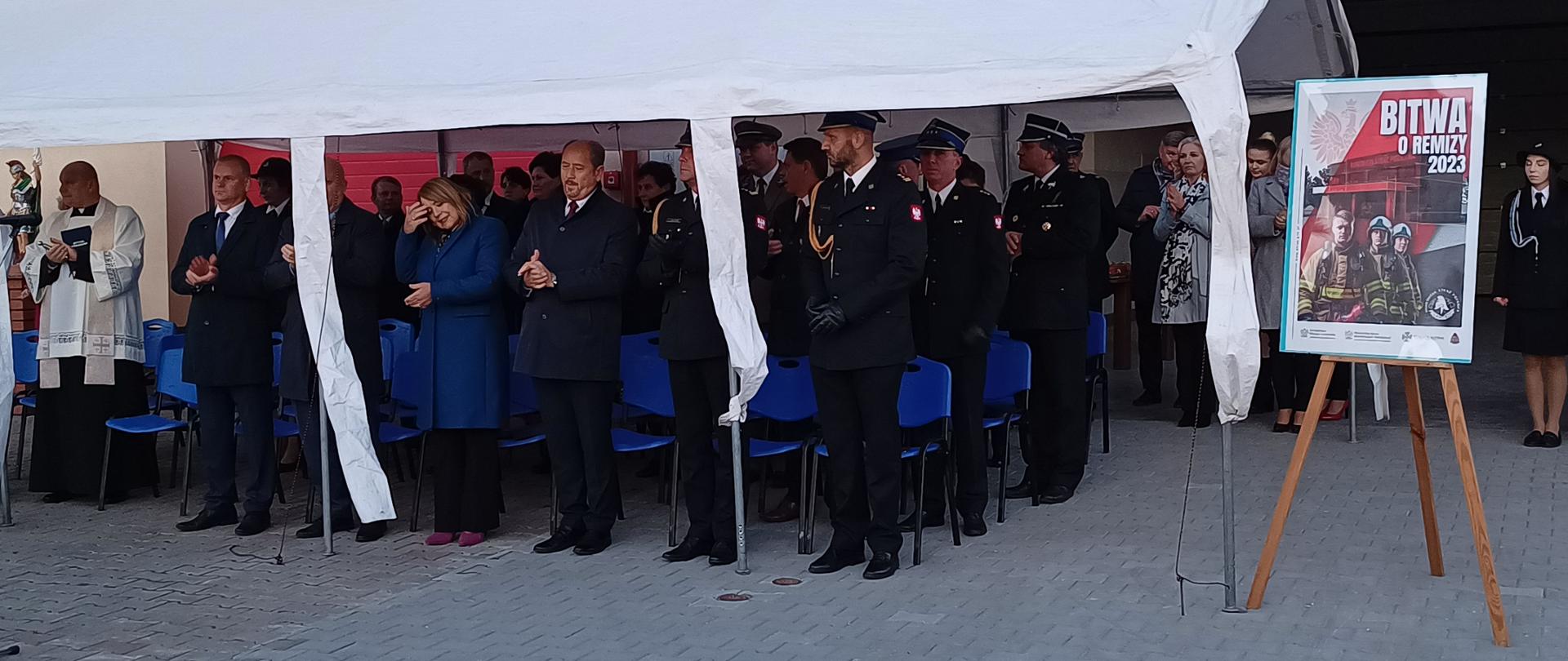Na zdjęciu widać gości zgromadzonych pod namiotem, przed budynkiem remizy strażackiej z prawej strony widoczny plakat.