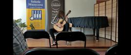 Dziewczynka z długimi ciemnymi włosami w bluzce w biało-czarne pasy i w czarnych spodniach siedzi na krześle i gra na gitarze jasnego koloru. W tle widać banery reklamowe Szkoła Muzyczna I stopnia w Tuchowie i Quena świat instrumentów. Po prawej stronie za grającą stoi czarny fortepian. 