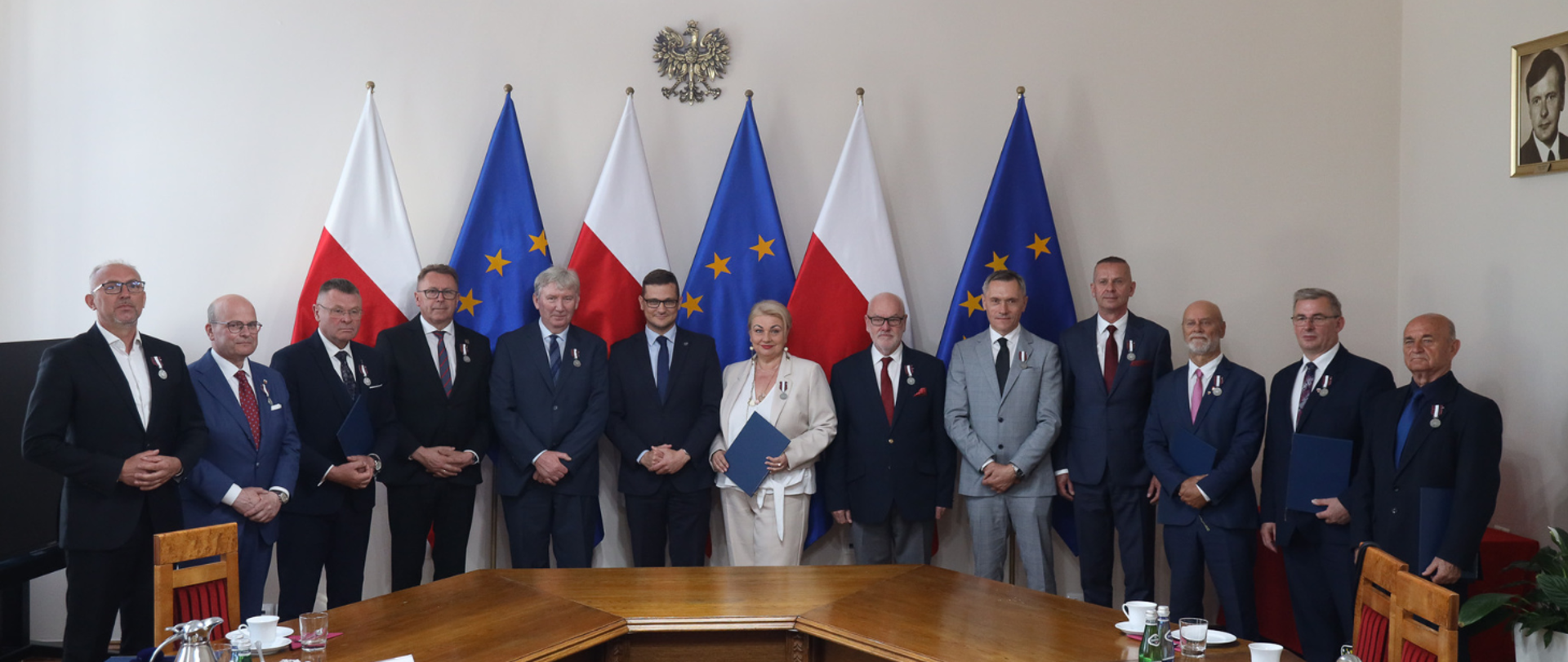 Dwanaście odznaczonych osób pozuje w rzędzie do zdjęcia z Wojewodą Kujawsko-Pomorskim Michałem Sztyblem na tle flag Polski i Unii Europejskiej oraz godła naszego kraju. 
