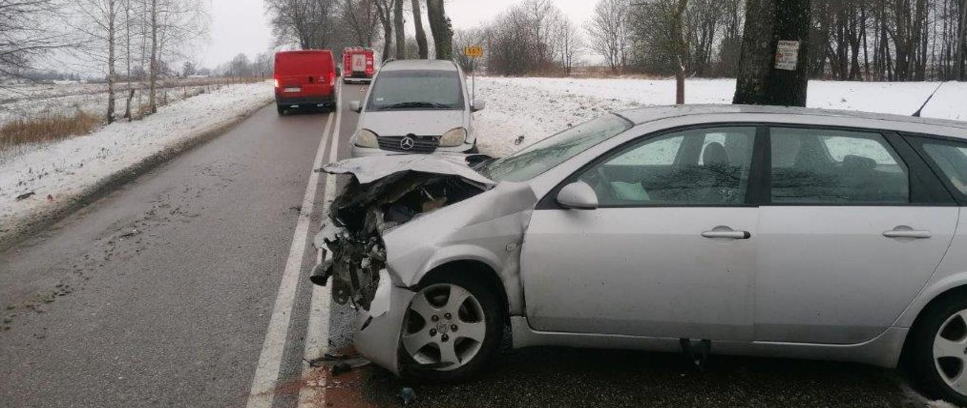 Zdjęcie przedstawia dwa samochody osobowe po zderzeniu. Jeden z nich ma uszkodzony przód i ustawiony jest prostopadle do drogi. Drugi stoi na prawym pasie ruchu bez widocznych większych uszkodzeń. W oddali stoi samochód pożarniczy. Ruch odbywa się wahadłowo. Na lewym pasie przejeżdża samochód.