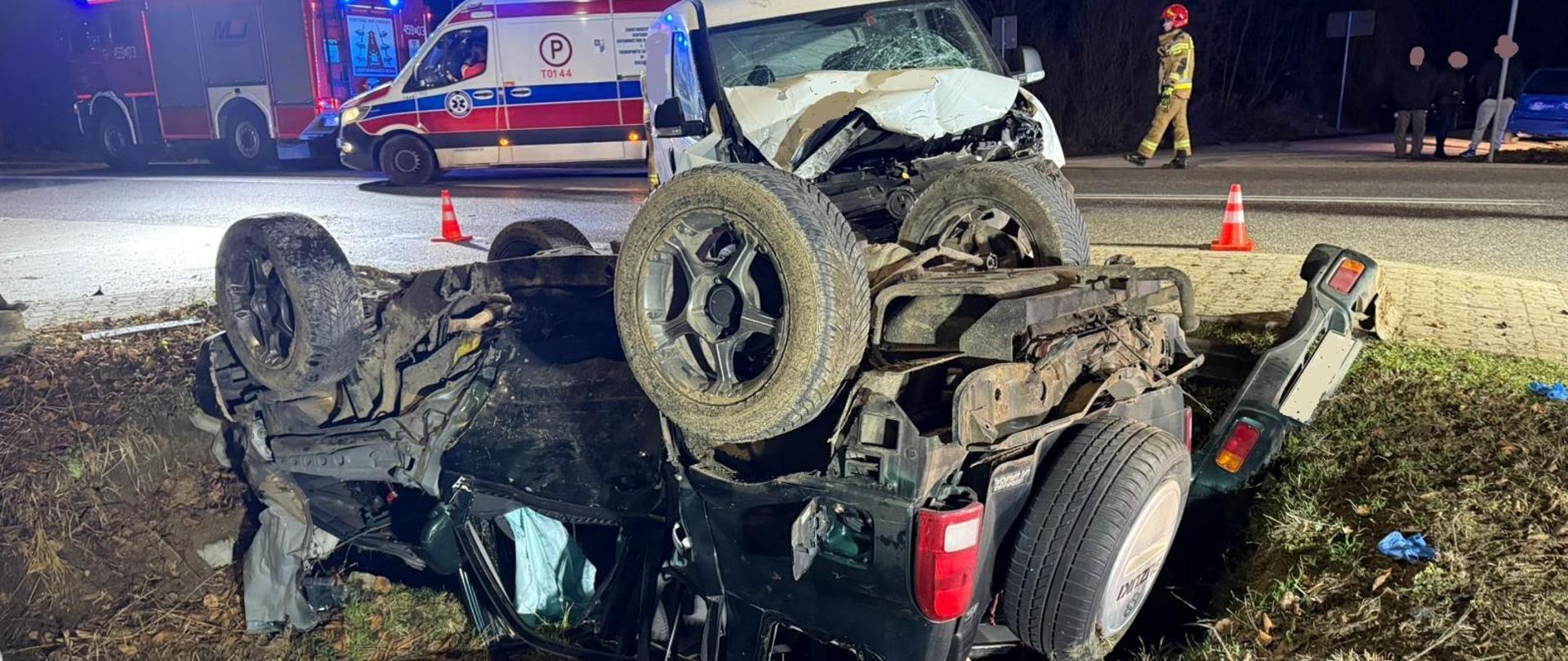 Na zdjęciu na pierwszym planie widoczne są samochody osobowe po zderzeniu drogowym. Jeden z nich znajduje się na dachu w przydrożnym rowie. Na drugim planie widoczny jest samochód zespołu ratownictwa medycznego oraz ratowniczo-gaśniczy.