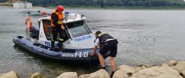 Widok z przodu. Łódź motorowa Policji przy brzegu. Na łodzi widoczni dwaj strażacy PSP. Na brzegu, widoczny od tyłu, policjant odpychający łódź od brzegu. W tle widoczny fragment zapory wodnej.