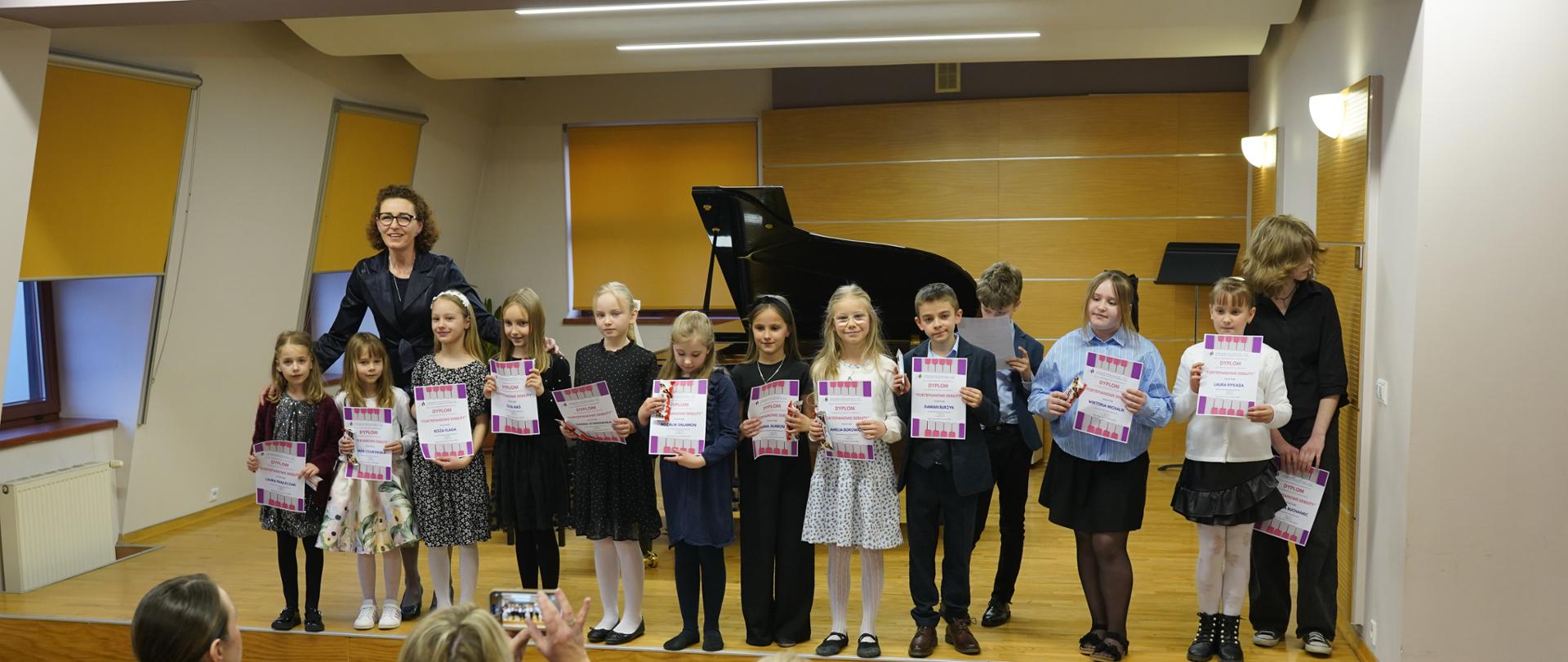 Zdjęcie przedstawia grupę dzieci stojących na scenie w sali koncertowej lub szkolnej, prawdopodobnie po występie muzycznym. Pierwszy plan: Grupa dzieci trzyma pamiątkowe dyplomy. Są ubrane odświętnie, a obok nich (po lewej stronie) stoi uśmiechnięta dorosła kobieta w okularach, prawdopodobnie nauczycielka lub organizatorka. Tło: W centralnej części sceny znajduje się czarny fortepian. Ściany są wyłożone jasnym drewnem, a oświetlenie jest ciepłe i punktowe. Perspektywa: Zdjęcie jest robione z poziomu widowni. Na dole kadru widać plecy osób siedzących w rzędach, które fotografują dzieci telefonami komórkowymi. Całość ma radosny, uroczysty charakter związany z sukcesem młodych wykonawców.