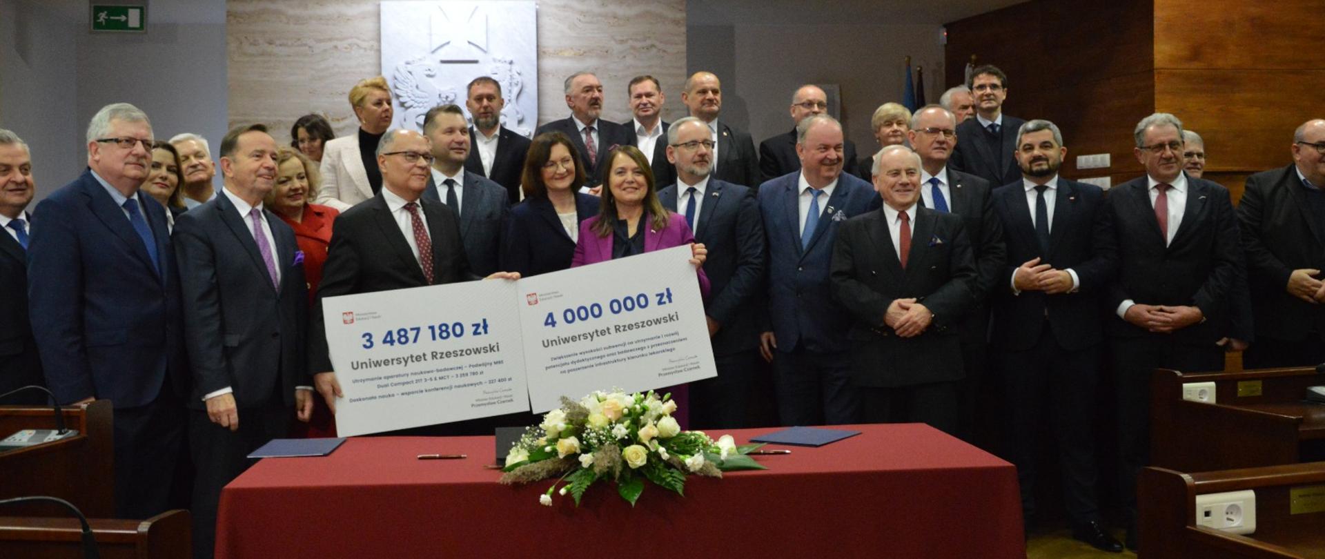 Duża grupa ludzi stoi przy czerwonym stole, kilka osób z przodu trzyma dwa duże symboliczne czeki z napisami 3 487180 zł i 4 000 000 zł.