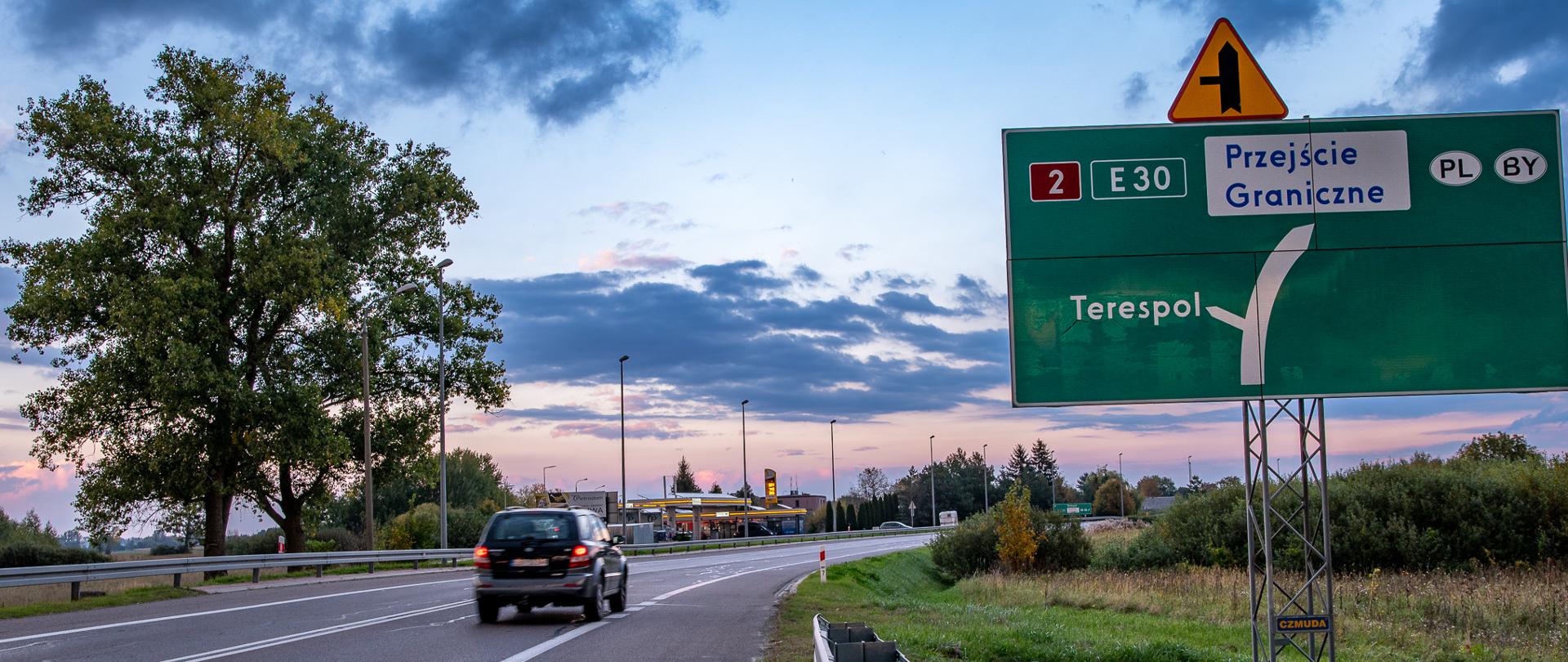 Zdjęcie przedstawia jezdnię i samochód. W tle widać stację benzynową. Po prawej stronie widać dużą, zieloną tablicę wskazującą drogę na Terespol oraz na Przejście Graniczne.